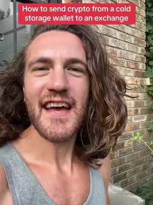 A man with long, wavy brown hair and a beard is smiling widely at the camera. He is wearing a gray tank top, and there's a red banner at the top with white text. Behind him is a brick wall and some greenery.