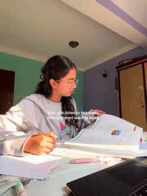 A young woman with long dark hair and glasses is seated at a desk, engrossed in studying. She's wearing a pale hoodie and is holding a pen over a paper. The desk is cluttered with open books, papers, and a laptop. The backdrop includes a colorful room with a large wooden cabinet. The overall lighting is soft, and the setting evokes a study environment.
