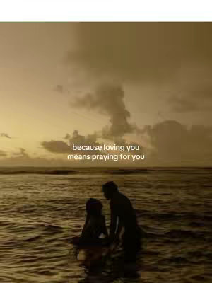 Two silhouetted figures, appearing to be a couple, stand in the ocean at sunset. The sky is a warm, muted yellow with some cloud coverage. Text overlaid on the image says, "because loving you means praying for you."