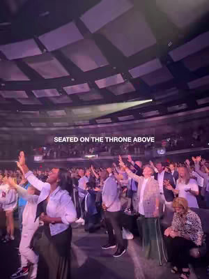 An image taken from slightly below, capturing a diverse crowd in a large venue with a high ceiling. Many people have their hands raised, presumably in worship, in purple and white lighting. At the top, the text 'SEATED ON THE THRONE ABOVE' overlays the shot.