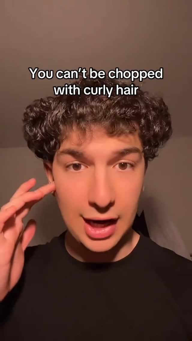 A close-up shot of a young man with dark curly hair. The background is a solid light brown. The man is looking directly at the camera with a slight smile, and his right hand is gesturing near his left ear.