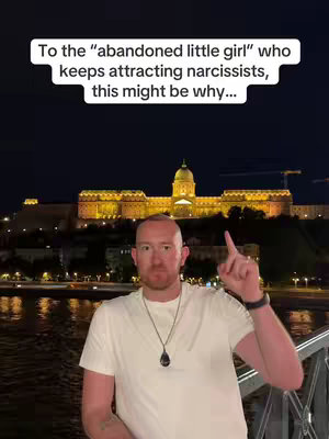 A man stands in front of a brightly lit old building at night near a body of water. He is wearing a white t-shirt and pointing upwards. Text overlaying the image reads: 'To the \"abandoned little girl\" who keeps attracting narcissists, this might be why...'