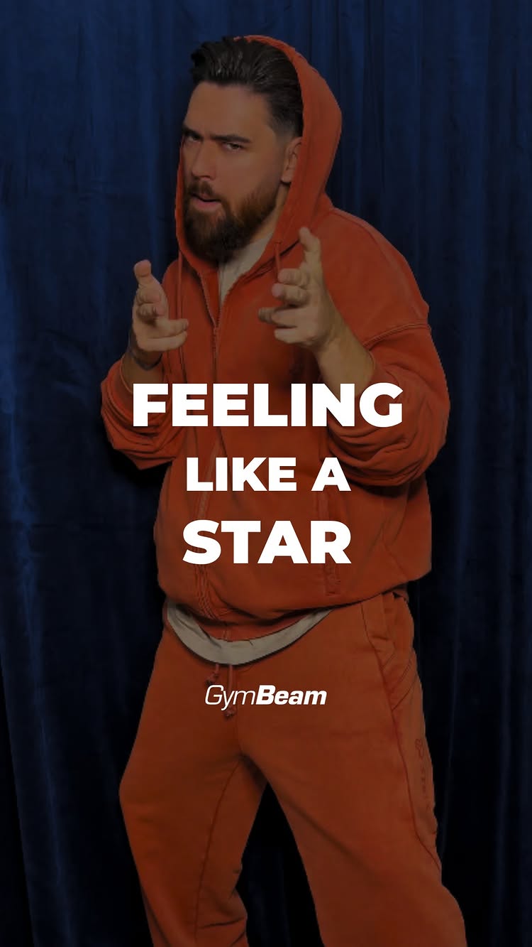 A man in an orange hoodie and track pants is posing in front of a dark blue curtain. The text overlay reads "FEELING LIKE A STAR". The GymBeam logo is visible at the bottom.