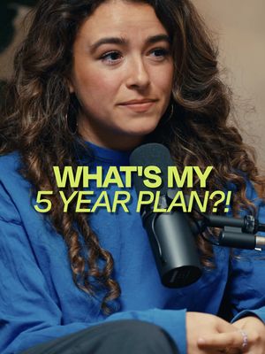 A medium shot features a woman with curly brown hair and fair skin, facing slightly to the left with a neutral expression. She wears a blue sweatshirt and has gold hoop earrings. Behind her, a dimly lit background is visible. Overlayed on the image, large yellow text reads, 'WHAT'S MY 5 YEAR PLAN?!' Positioned near her face are a microphone and stand.