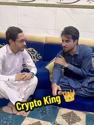 Two men are seated on a blue couch, engaged in conversation. The man on the left wears glasses and white attire, and watches the man on the right, who is wearing a blue outfit and has an animated crown near him, suggesting leadership or expertise, perhaps dealing with crypto.