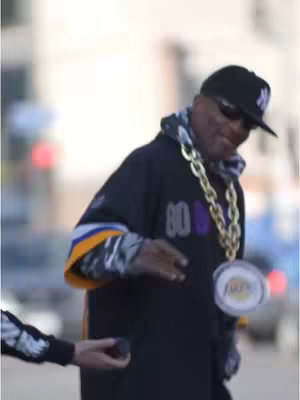 The image captures a man in a casual setting, possibly outdoors. He's wearing a black baseball cap with a New York logo, sunglasses, and a black jersey with the number 80 and white and purple accents. A large gold chain with a Lakers pendant adorns his neck. A hand holding a dark object is partially visible on his left. The background appears blurred, suggesting a shallow depth of field.