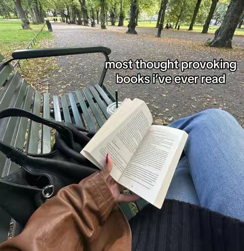 A first-person view of a person reading a book outdoors. The person is sitting on a park bench. The open book is in the foreground and has pages with text. The background is a park walkway with trees, and leaves.