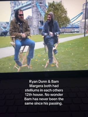 A shot of two men sitting in director's chairs in front of the Tower Bridge. The men, one with a beard and sunglasses, and the other in a hoodie, face each other. Text is superimposed at the bottom of the image.