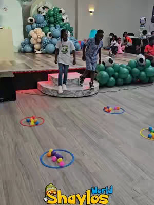 The cover image shows an indoor scene of a children's birthday party. Two children are walking down some white steps, there is a backdrop created with blue and white balloons to the left of the children, and a series of brightly-colored toys and hoops on the floor. A logo is present in the foreground, and there are additional children and adults in the back. Soft lighting, and the scene suggests a fun environment.