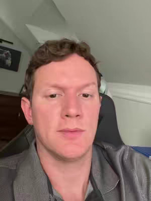 A medium shot of a man facing the camera, indoors. The room appears to be a home office or bedroom, with a framed picture on the wall and a black chair visible. The man has short, dark, curly hair and is wearing a casual gray shirt. His facial expression is neutral, looking directly at the viewer.