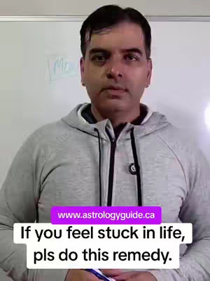 A medium shot shows a man in a gray hooded sweatshirt in front of a white board. He is looking directly at the camera. Text overlayed at the bottom says 'If you feel stuck in life, pls do this remedy.' Above that, a website listed as www.astrologyguide.ca. The background has a whiteboard with some text written on it. The man has short, dark hair.