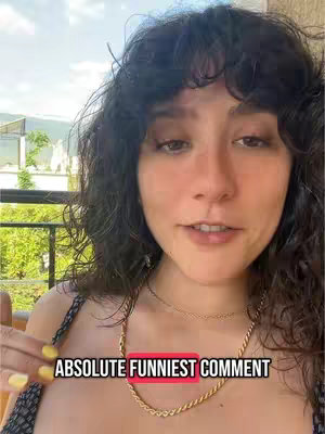 A woman with dark, curly hair smiles at the camera in a selfie style, with a red rectangular overlay and text at the bottom that reads 'ABSOLUTE FUNNIEST COMMENT'. She wears a black top, and a background of trees and a balcony is visible. The lighting appears to be natural daylight.