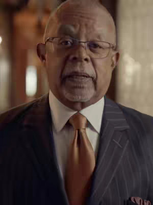 An older, fair-skinned man with short, graying hair and glasses is centered in the frame, looking directly at the viewer. He wears a dark pinstripe suit with a white shirt and a gold tie. The background is slightly blurred, suggesting an indoor setting.