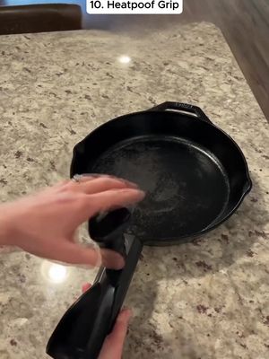 A close-up shot of a kitchen countertop with a cast-iron skillet. A hand wearing a ring holds a black heatproof grip, appearing to apply it to the skillet's handle. The countertop is a light, speckled color.