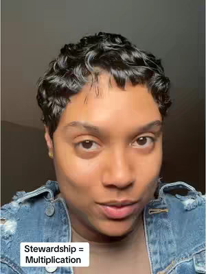 A close-up shot of a person with short, wavy black hair. The person is looking slightly to the side, with a neutral expression. They are wearing a denim jacket. In the bottom left corner, there is white text that says 'Stewardship = Multiplication'. The background is a soft, blurred shade of grey.