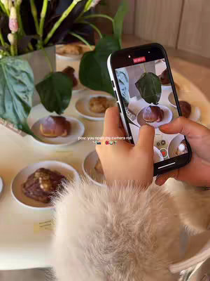 A person is holding a smartphone in their hands. The phone's screen displays a close-up of a dish with a pastry. The background shows a softly lit cafe with plates of similar pastries on a table and some plants.