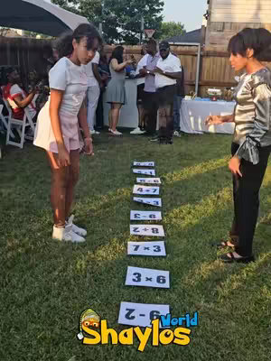 Two young women stand on a grassy lawn, facing each other across a series of numbered rectangular cards arranged in a line. The background shows a casual outdoor gathering with a gazebo and other people.