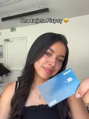 A young woman with long dark hair holds up a blue credit card in front of a white wall and overhead lights.