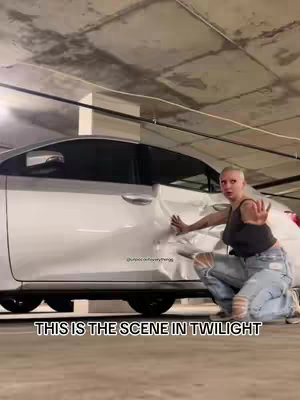 A woman in ripped jeans and a tank top kneels next to a white car in an underground parking garage. The car has a dent in the side. She extends her hand towards the car, as if stopping something.