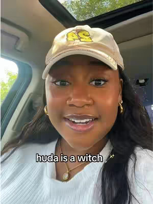 A woman in a car wears a baseball cap and jewelry, looking directly at the camera and smiling. Text overlay says "huda is a witch".