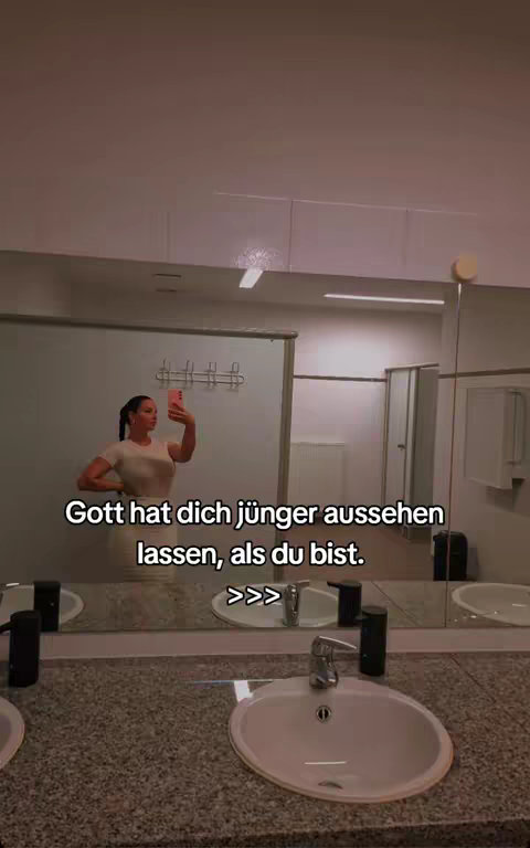 A woman is in a bathroom, taking a selfie in front of a mirror. The mirror displays a reflection of the space, including a sink, various toiletries, and the woman. Text overlayed on the image reads, "Gott hat dich jünger aussehen lassen, als du bist. >>>".