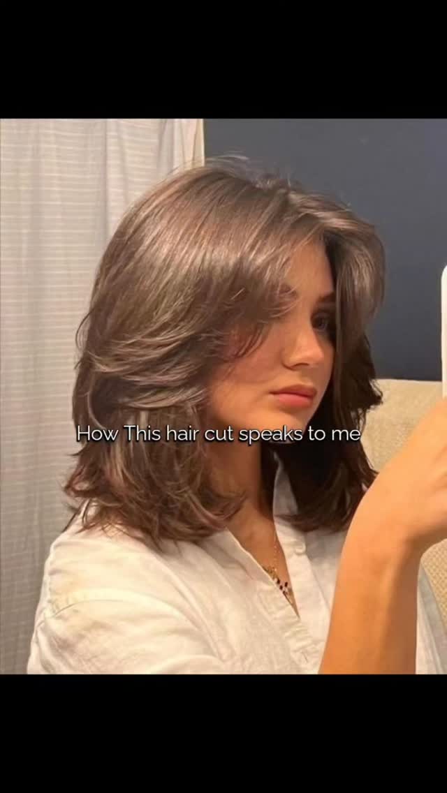 A young woman with medium-length, layered brown hair, styled in a modern haircut. The woman is fair-skinned, wearing a white button-down shirt, and seems to be taking a picture of herself. The background includes a curtained window and a dark-colored wall. A text overlay is written over the image.