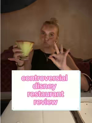 A young woman with blonde hair in a bun holds a yellow drink and gestures an ok sign. Text overlayed says: 'controversial disney restaurant review.' The background is a blurred interior.