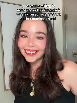 A smiling woman with brown hair and fair skin is the focal point of the image. She is positioned in front of a light-colored wall, possibly a bathroom. Above her, text overlays read "pov: you're talking to someone and noticing she's wearing an evil eye bracelet on her dominant hand". The woman's expression is friendly, and she has rosy cheeks and red lipstick. A necklace with charms hangs around her neck. The lighting appears to be natural, and the overall composition is simple, focusing on the woman's face and the overlaid text.
