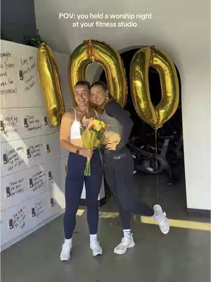 Two women stand in front of a wall with lockers and a large gold '100' balloon display in a cycling studio. The woman on the left holds a bouquet of flowers, and both are smiling. The setting indicates a celebration or event within a fitness context.