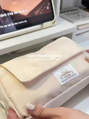 The image shows a close-up of a woman's hands holding a beige pencil case. The pencil case has a flap, and a small white tag with a cartoon character illustration and text is visible. Behind the pencil case, a blurred image of a tablet is visible, along with a white shelf and a book. The overall composition is clean and minimalist.
