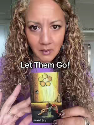 A woman with curly auburn hair and a serious expression holds a tarot card in front of her. The card depicts images of coins and a door. The photo is framed from the chest up with the woman wearing a purple top. The background appears to be a room with white cabinets.