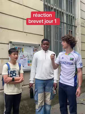Three young men standing outside a building. The central figure is wearing a white shirt and jeans with patchwork pockets. The other two are on either side, and one is wearing a soccer jersey. A red banner in the image reads "réaction brevet jour 1".