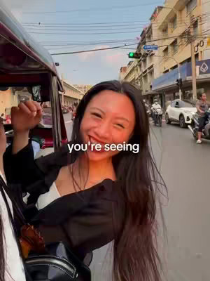 A woman with long, dark hair smiles broadly while riding in a tuk-tuk. The vehicle is moving along a city street, with buildings and traffic in the background. Overlay text says: "you're seeing".