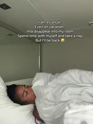 A person lies in bed, mostly covered by a white comforter. The person's face is visible, with closed eyes, suggesting sleep or relaxation. Text is overlaid on the image, stating 'I am a cancer. Even on vacation, Ima disappear into my room. Spend time with myself and take a nap. But I'll be back.'
