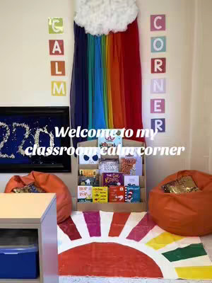 The image shows a classroom corner decorated for a calming area. A rainbow-colored fabric hangs from the ceiling, resembling a rainbow. Books are displayed on a bookshelf. Two orange beanbag chairs are placed on either side of the bookshelf. On the floor, there is a painted sun design. On the wall, there are letters spelling out "CALM CORNER".