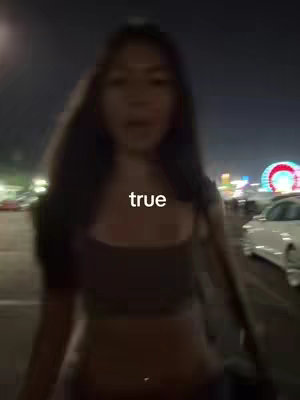 A woman is filmed walking towards the camera at night. The background is blurred and features city lights, including a Ferris wheel. Text overlayed on the video says 'true.' The woman is wearing a brown top, and has long dark hair. The shot is slightly out-of-focus.