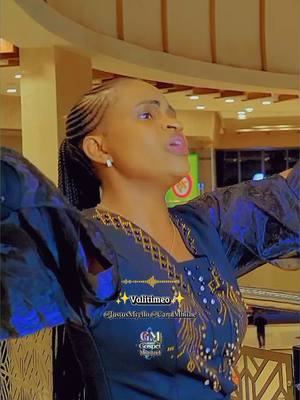 A woman with braided hair in a dark, patterned dress, arms outstretched. The background is an interior space with warm lighting.