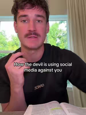 A medium shot features a man looking directly at the camera. He has short, dark hair, a mustache, and is wearing a black t-shirt with the word "GRACE" on it. The text overlay reads, "How the devil is using social media against you". Behind him, a window is visible, with outdoor scenery, including green plants and foliage.
