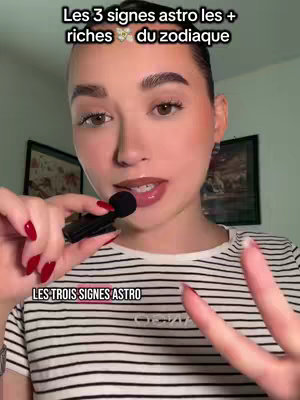 A woman with light skin stands in front of a background with pictures and a green wall, holding a small microphone with the index and middle fingers of her right hand raised in a 'peace' gesture. She appears to be smiling slightly, and wearing a black and white striped shirt with red nail polish on her fingers.