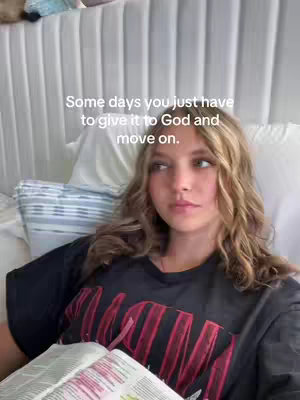 A young woman with long, wavy blonde hair is lying in bed, looking slightly off-camera. The background shows a white, textured headboard and a blue-and-white striped pillow. She's wearing a black t-shirt with a red design, and she is holding a book with visible text which is open. The scene has soft, natural lighting, creating a relaxed and intimate atmosphere.