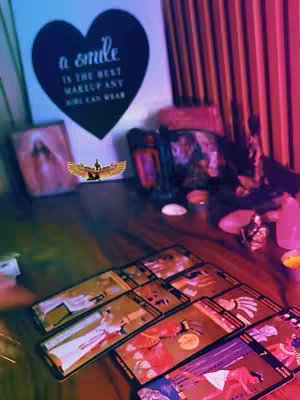 The image is a well-lit, aesthetically arranged setup featuring a wooden surface covered with tarot cards, crystals, and various spiritual objects. The composition draws the eye with a balanced distribution of elements, suggesting a reading or ritualistic context. A heart-shaped artwork saying 'a smile is the best makeup any girl can wear' is visible in the background, as well as other symbolic images.