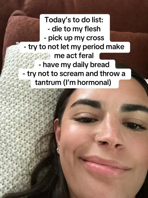 The cover image showcases a young woman in a close-up shot, lying down with her head resting on a light-colored knitted surface, likely a pillow or cushion. The woman has dark hair and is looking at the camera with a slight smile. The background consists of soft shades of brown, likely a couch or bedding, complementing the overall warm tone. A text overlay, presented in a simple, sans-serif font, is layered at the top, listing items for the day in a humorously exasperated tone.