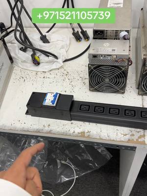 A person's hand points to a black power distribution unit (PDU) with multiple electrical outlets. Various electronic components, cables, and a silver power supply unit are visible in the background.