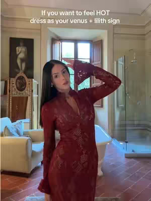 A woman in a long-sleeved, red lace dress poses in a well-lit, elegant room. The background includes a large window, a white bathtub, and decorative furniture.