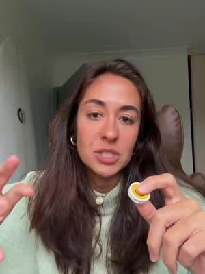 A woman with long, dark hair and a light green sweater is the primary focus, set against a warmly lit indoor setting. She is holding a small, round object with a smiley face, and is gesturing with her other hand. The background includes a glimpse of a brownish-colored chair and wall decor.