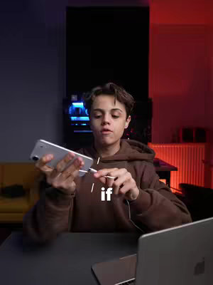 A young person in a brown hoodie is holding a silver iPhone and plugging in a charging cable. They are seated with a laptop in front of them. The background has a sofa and a computer screen on a desk.