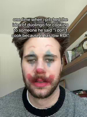 A man with clown makeup on his face looking into the camera. The background has a wooden shelf with books and other items.