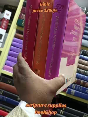 A hand holding a stack of books, the spines of which are visible. The books are in shades of red, orange, and purple, and the titles are partially visible. Behind the books are more books on shelves. In the foreground are the creator's hand.