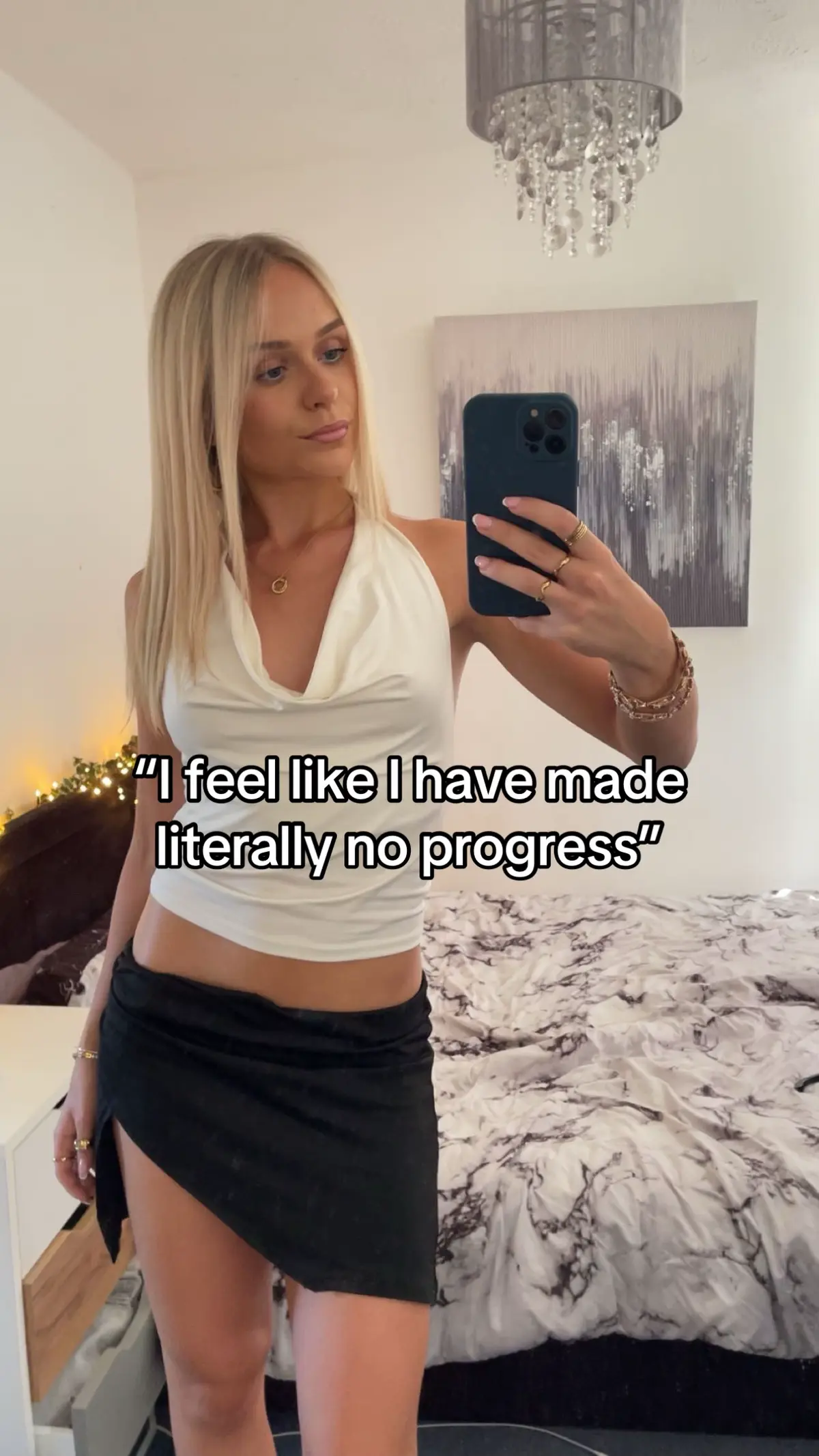 A young woman is taking a mirror selfie, she is wearing a white halter top and a black mini-skirt. She is holding a phone up with her right hand, the phone is covering her face. A reflection of the room is visible in the mirror behind her, with a painting and a chandelier on the top, a bed on the bottom.