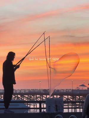 A silhouette of a person blowing a large bubble with an orange and pink sunset in the background. The bubble is in the shape of a heart. The person is standing on a concrete surface next to a white bucket, with a pier and ocean visible in the background.